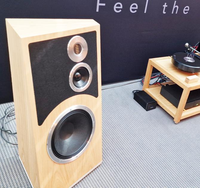 The Audiovector Trapeze speaker in Oak with a low unit just visible to the right that has other audio equipment on it.