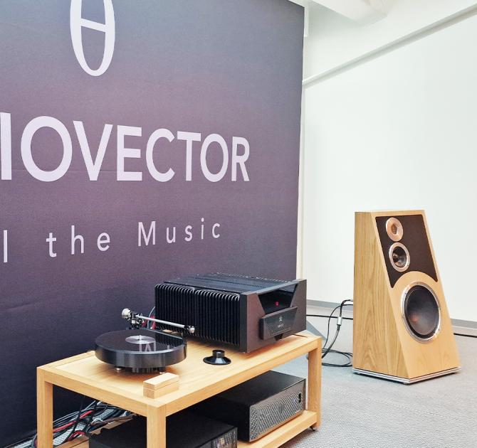 The Audiovector Trapeze speaker in Oak with a low unit to the left that has other audio equipment on it.