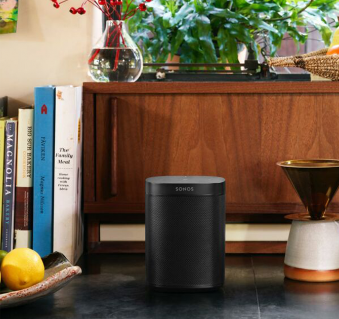 Sonos One Black next to a bowl with a lemon in with a wooden sideboard behind it with cookery books stood up to the left and a vase on top with stems of red berries in.