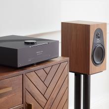 Linn 119 Loudspeaker in walnut on the black stand beside a wooden sideboard with a DSM on top of it.