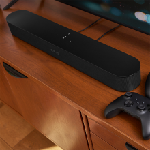 SONOS Beam (Gen 2) on a TV cabinet with a gaming controller beside it.  The bottom of a flat-screen tv is visible.