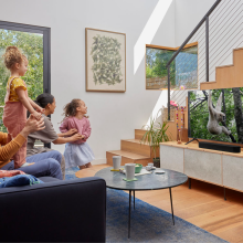 SONOS Beam (Gen 2) in black on a tv cabinet in front of a flat-screen tv which has a monkey on it.  there are stairs to the side of the tv unit.  there's a coffee table in front of the tv unit and a family sitting.  Two children are playing - one is standing up on her dad's legs.  he's seated.