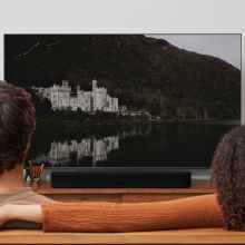 SONOS Beam (Gen 2) in black.  there's a man in the foreground on the left and a woman on the right.  She has her arm on the back of the sofa.  the tv is showing a picture of a castle reflected in a lake with a hill behind it.
