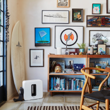 SONOS Sub in white on the floor in front of a surf board, a wall with lots of framed pictures and a cluttered shelving unit.