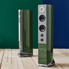 A pair of tall, slim speakers in a green colour on a wooden floor in front of a dark green wall