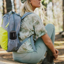 SONOS Roam SL in black in the side pocket of a backpack.  The woman is sitting on the floor.