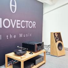 The Audiovector Trapeze speaker in Oak with a low unit to the left that has other audio equipment on it.