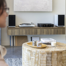 A pair of Moon Voice 22 Loudspeakers in white with grille on standing on a wooden sideboard.  There's a low wooden table in the middle of the image and a woman in the foreground.