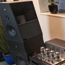 A Rosso Fiorentino Volterra speaker in black with a Synthesis Roma 510AC amplifier on the bottom right of the image
